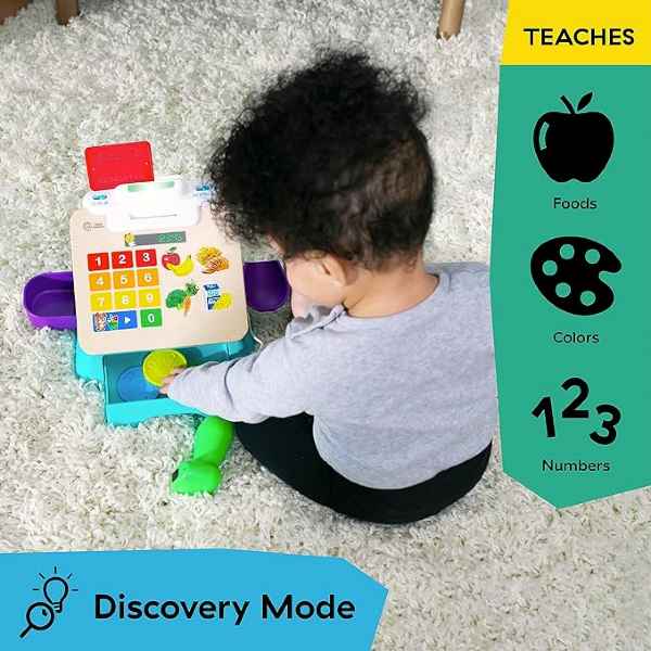 Toddler playing with a colorful wooden toy register featuring a numeric keypad and food icons on a white plush rug.