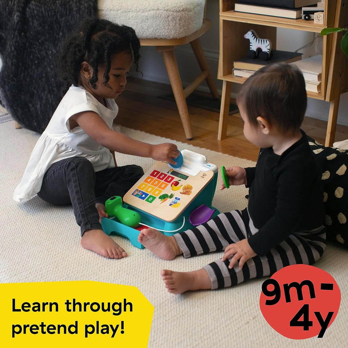 Two toddlers play with a teal and wood toy cash register featuring colorful number buttons and a green hand scanner.