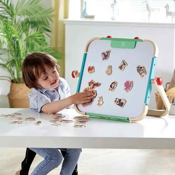 Child placing wooden animal magnets on a white magnetic easel with a light wood frame at a white table.