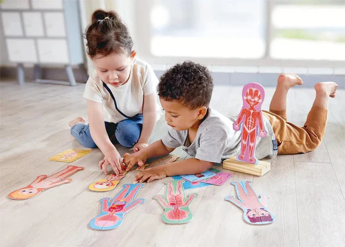 Two children play with wooden puzzle pieces depicting the human skeletal, muscular, and organ systems on a light wood floor.