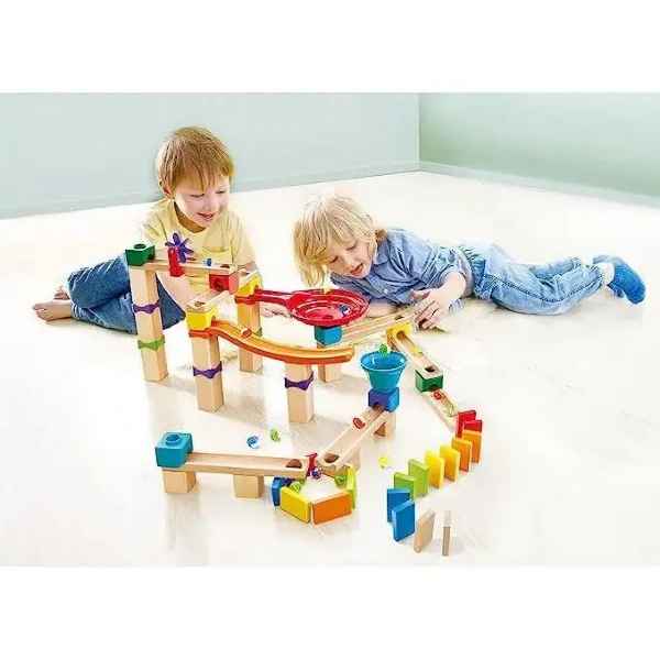 Two children playing with a wooden marble run with colorful blocks, a red funnel, and dominoes on a white floor.