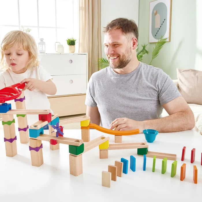 Man and child build a wooden marble run with colorful connectors, ramps, and dominoes on a white table.