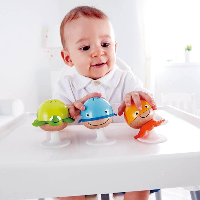 Baby plays with three animal-themed suction rattles: a green turtle, blue whale, and orange octopus on a white tray.