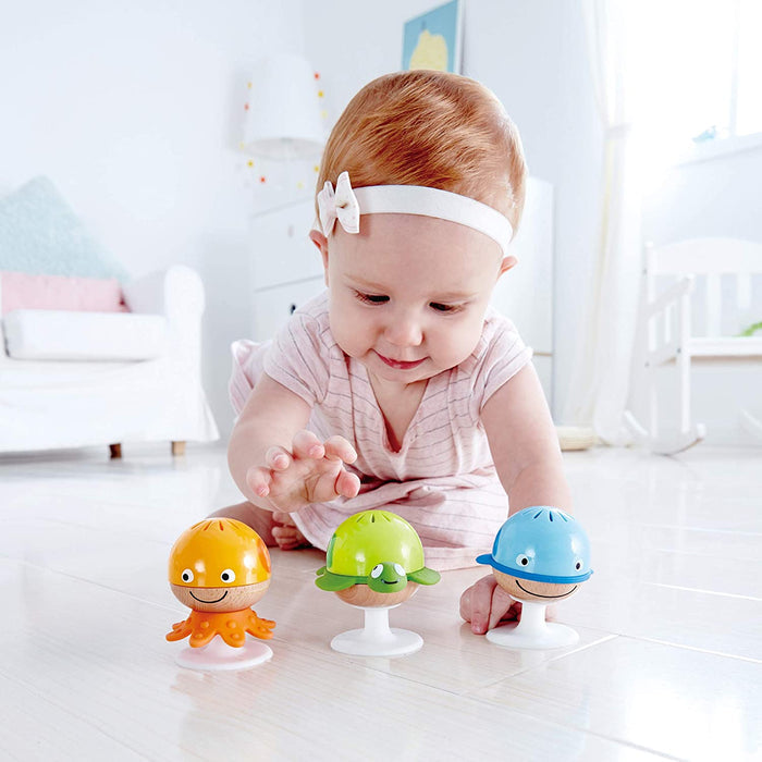 Baby playing with orange octopus, green turtle, and blue whale suction cup rattles on a white floor.