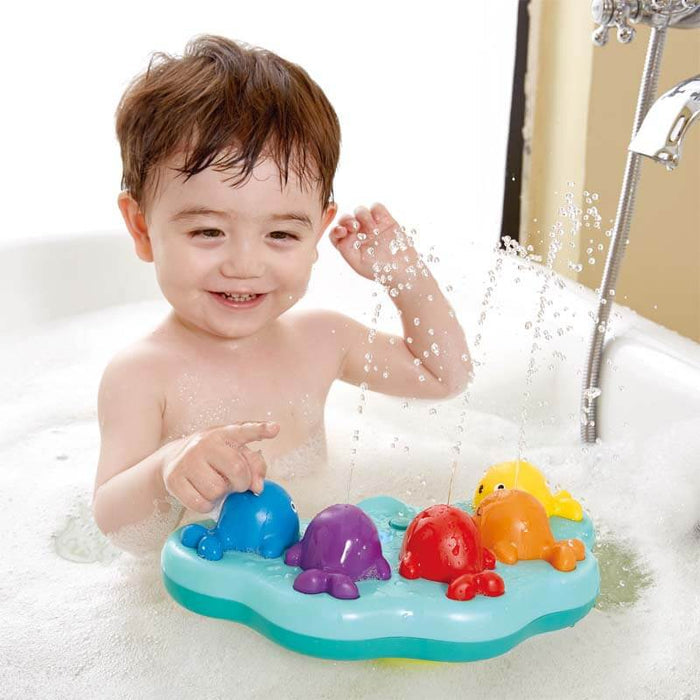 Toddler smiles as five colorful toy whales on a light blue floating base spray water in a bubbly bathtub.
