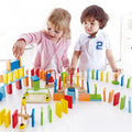 Two children build a colorful wooden domino course with bridges, ramps, and bells on a bright white table.