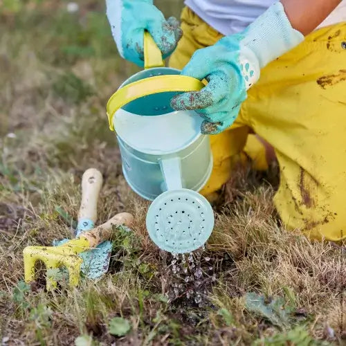 Child in yellow pants pouring a light blue watering can near a yellow hand rake and trowel on a patch of grass.