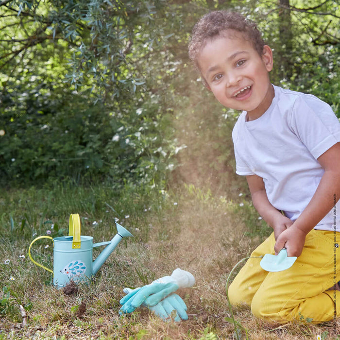 Smiling child in a garden with a light blue hedgehog watering can, matching gloves, and a small blue spade.