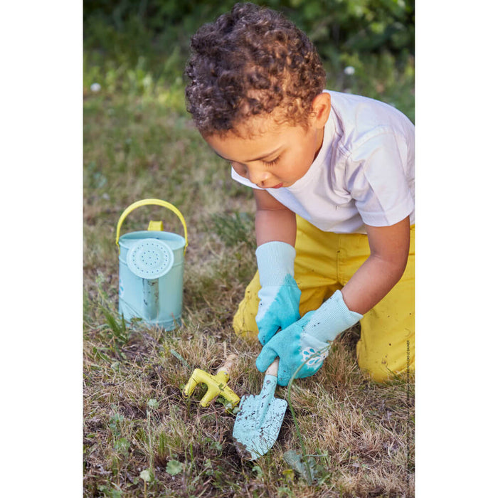 Child in gardening gloves uses a light blue hand shovel and yellow rake next to a matching watering can in a garden.