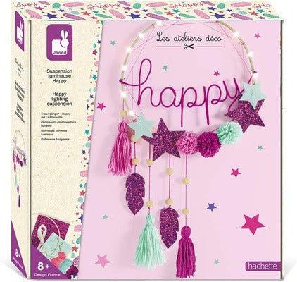 Pink craft kit box featuring a lighted hoop with pink 'happy' script, glitter stars, pom-poms, and colorful tassels.