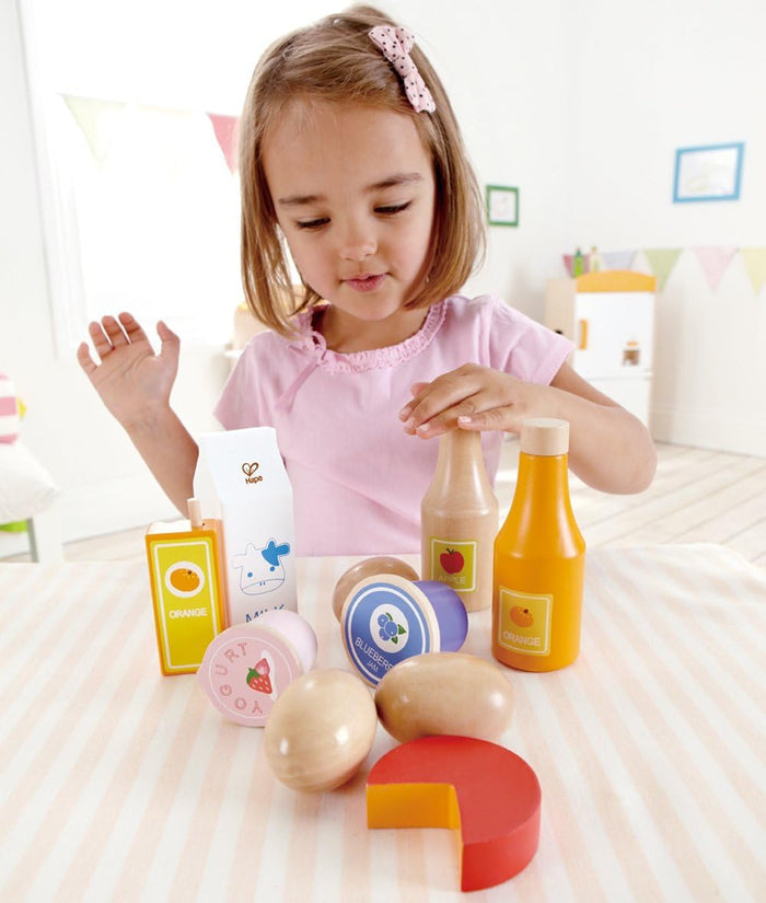 Young girl playing with colorful wooden play food: milk carton, juice bottles, eggs, and cheese on a striped tablecloth.