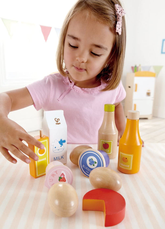 Young girl playing with colorful wooden toy food: milk carton, juice, jam, and natural wood eggs.