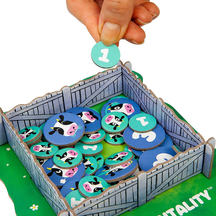 Hand dropping a teal number one token into a cardboard farm pen filled with circular cow face and number game tokens.