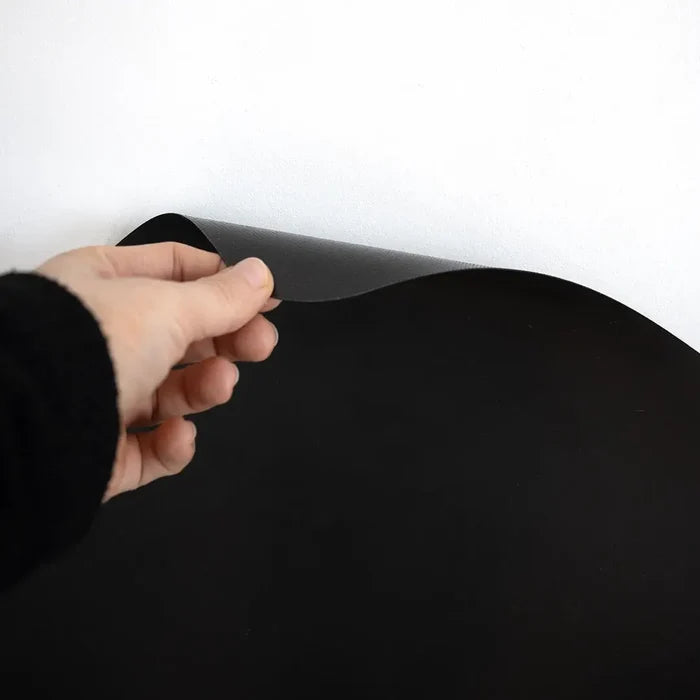 Hand peeling a thin, flexible black magnetic sheet against a white wall, showcasing the matte texture.