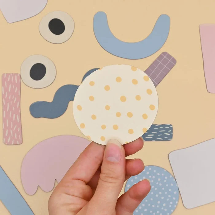 Hand holds a yellow polka-dot circle magnet over colorful abstract shapes like eyes and arcs on a beige board.