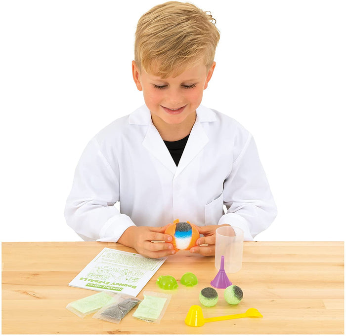 Boy in a lab coat making colorful bouncy eyeballs with plastic molds, powder packets, and tools on a wooden table.
