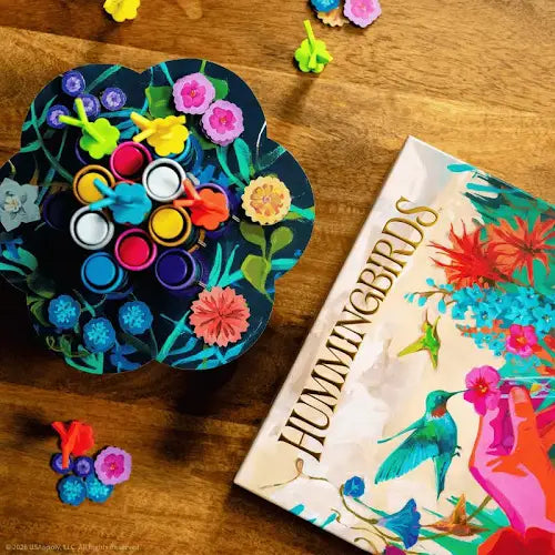 Flower-shaped game board with colorful pegs next to a Hummingbirds box with bird and floral illustrations.