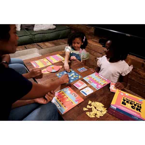 Two children play I Want My Teeth Back with pink boards and yellow tooth tokens next to the yellow game box.
