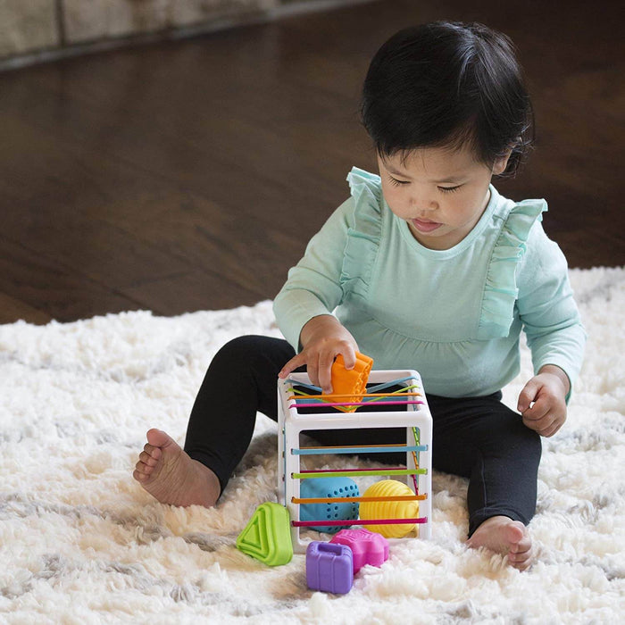 Baby playing with a white cube toy with colorful elastic bands and textured shapes on a soft white rug.