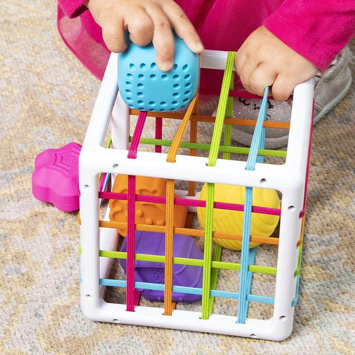 Child pushing a blue textured block through colorful elastic bands on a white sensory cube toy.