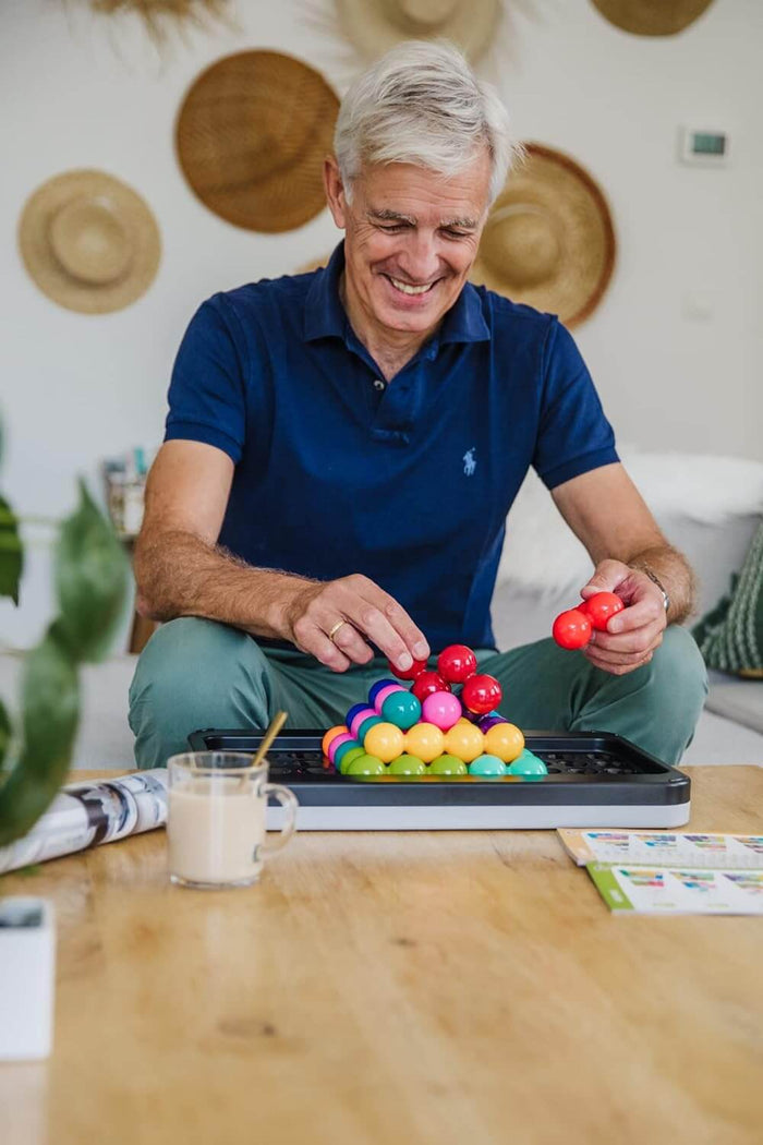 Smiling man builds a colorful 3D pyramid using connected sphere pieces on a large black and white game tray.