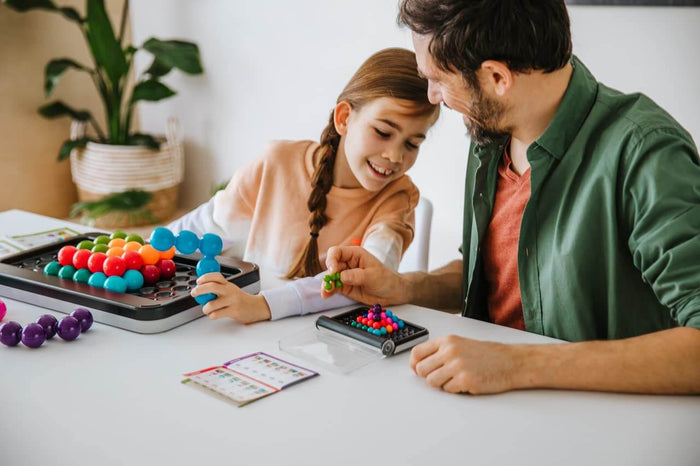 Man and girl solve colorful bead puzzles on a large black game board and a small portable case at a table.