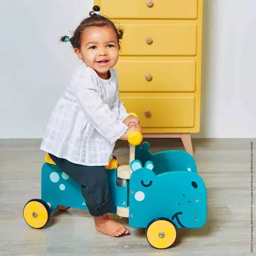 Toddler on a teal wooden hippo-shaped ride-on toy with yellow wheels, in front of a yellow dresser.