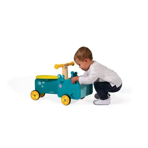 Teal wooden hippo ride-on toy with yellow wheels and seat being pushed by a small child on a white background.