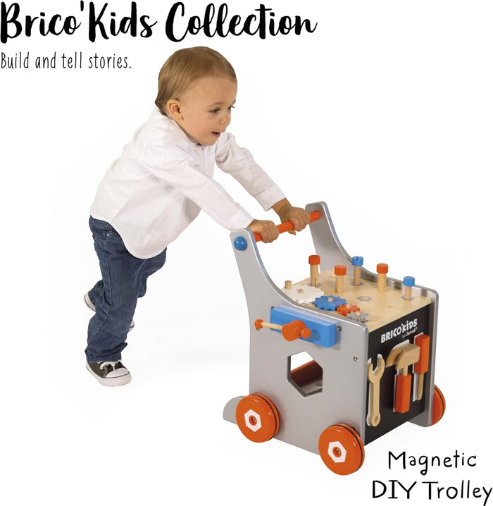 Toddler pushing a gray wooden trolley with orange wheels, workbench top, and magnetic tools attached to the side.