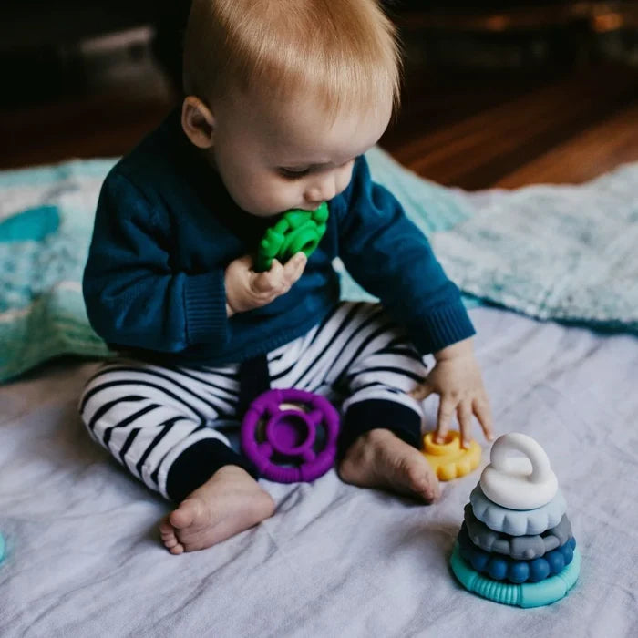 Baby chewing a green ring next to a stack of white, blue, and grey textured silicone rings on a blanket.