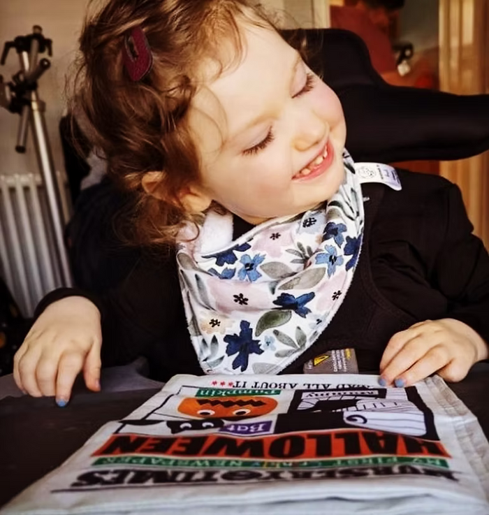 A young child wearing a black top and a white bib with blue and green floral patterns, smiling while looking down at a Halloween-themed printed sheet on a table.
