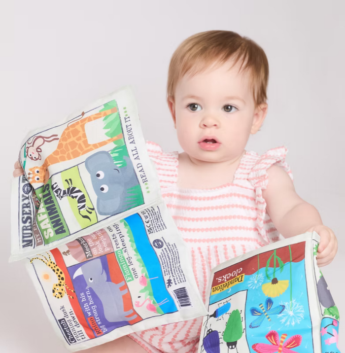 A baby girl in a pink and white striped dress holds two colorful children's picture books with cartoon animal and flower illustrations against a plain light gray studio background.