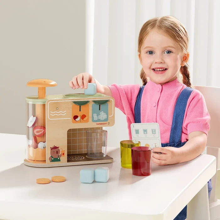 Smiling girl using a wooden juice dispenser toy with colorful cups, fruit slices, and blue ice cubes.