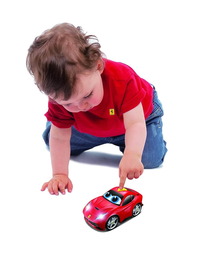 Toddler pressing a yellow button on a red Ferrari toy car with cartoon eyes and glowing headlights.