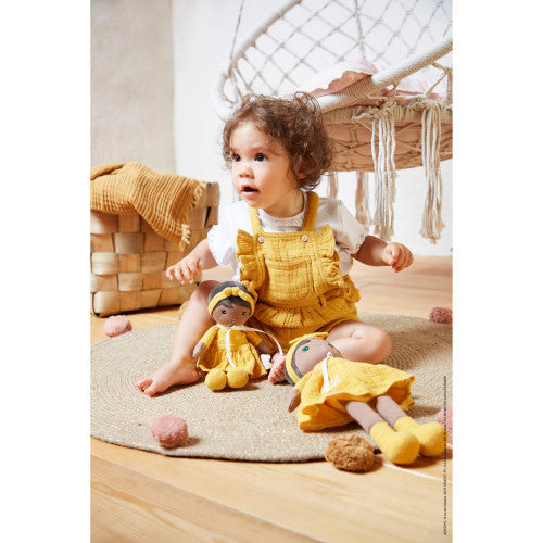 Baby playing on a rug with two soft cloth dolls wearing yellow dresses and headbands.