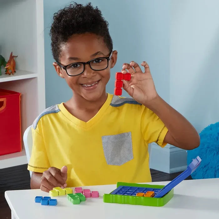 Smiling boy holding a red puzzle piece above a green game tray with colorful geometric blocks on a white table.