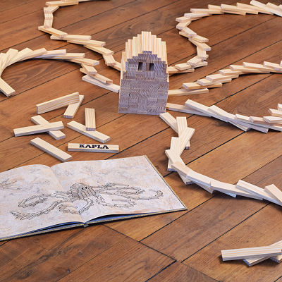 Open Kapla art book beside a wooden octopus sculpture built from beige planks on a hardwood floor.