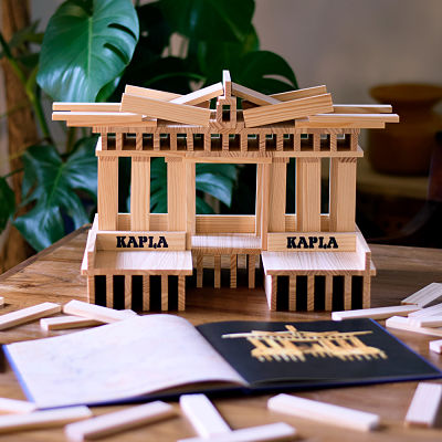 Wooden plank building next to the open blue Kapla Art Book showing an architectural model diagram on a table.