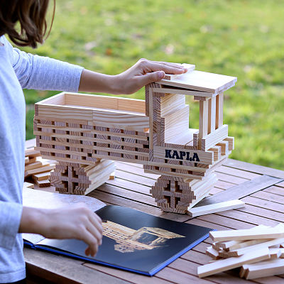 Open blue Kapla Art Book showing building designs next to a large truck constructed from wooden planks.