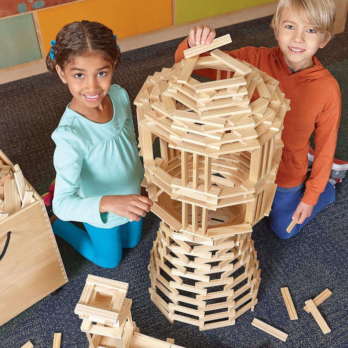 Two children build a tall, complex tower using light-colored wooden planks on a blue carpet next to a storage bin.