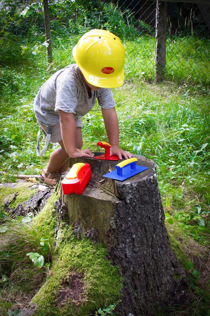 Child in a yellow toy hard hat plays with red and blue plastic tools on a tree stump in a grassy yard.