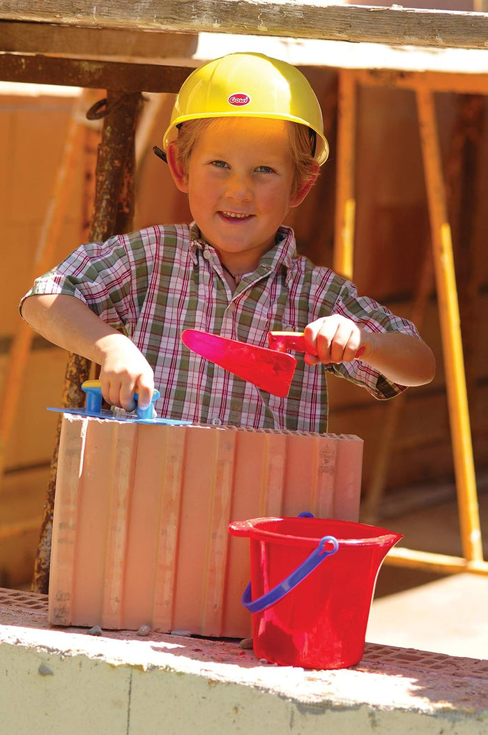 Child in yellow hard hat using red toy trowel and blue hawk on bricks, with a red toy bucket in the foreground.