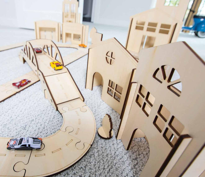 Light wood toy buildings with window cutouts and a wooden road track featuring a bridge and colorful toy cars.