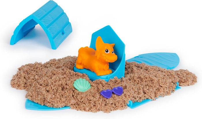 Orange toy dog on blue house parts in a pile of brown kinetic sand with a small shell and purple heart sunglasses.