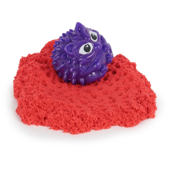 Purple spiky toy creature with large eyes sitting on a mound of textured red sand.