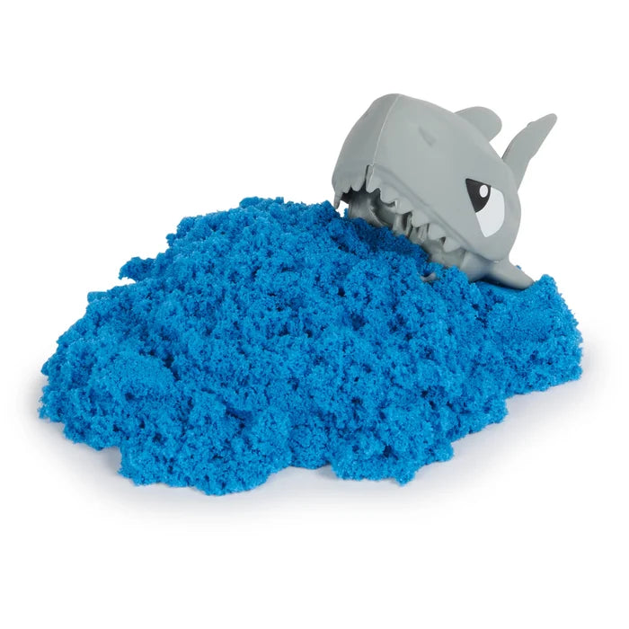 A pile of textured blue sand with a grey toy shark head popping out, showing white teeth and cartoon eyes.