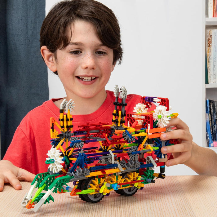 A boy smiles at a multi-colored toy vehicle with wheels and white gears, built from interlocking rods and connectors.