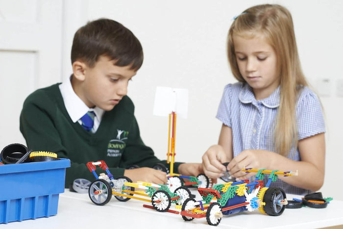 Two school children building colorful K'NEX cars with plastic rods and wheels on a white table.