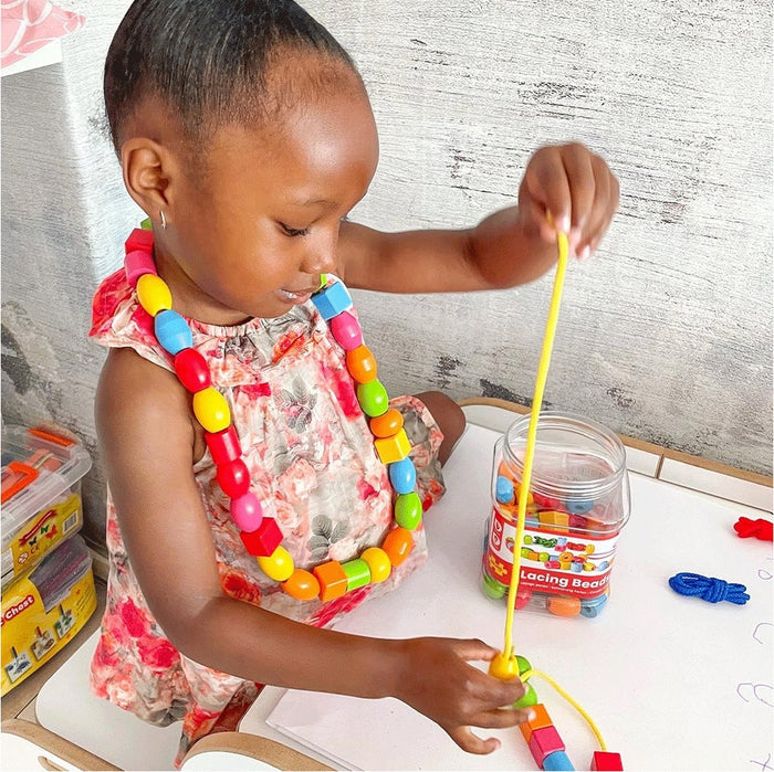 A child threads large, colorful wooden beads onto a yellow string. A clear jar of assorted beads sits on a white table.