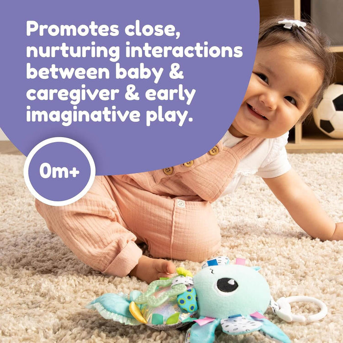 A baby in pink overalls smiles on a rug next to a colorful, plush axolotl toy with hanging textures and a white clip.